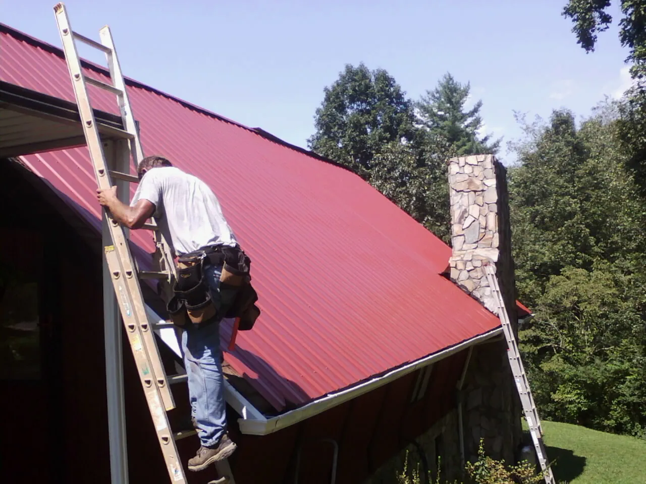 Roofer on a ladder beside a red metal roof and stone chimney — Soffit & Fascia Repair by Roofing Brothers in Reynoldsburg